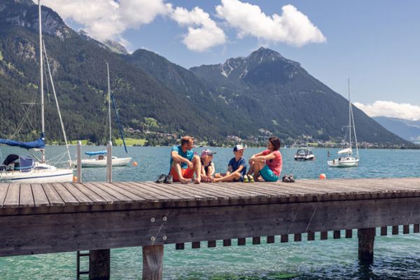 Familie sitzt auf einem Holzsteg an einem See, im Hintergrund Segelboote und Berge