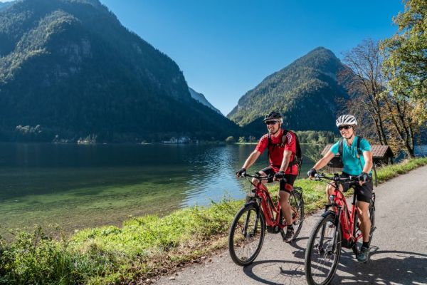 Eine Radfahrerin und ein Radfahrer fahren im Sommer an einem See, Berge im Hintergrund.