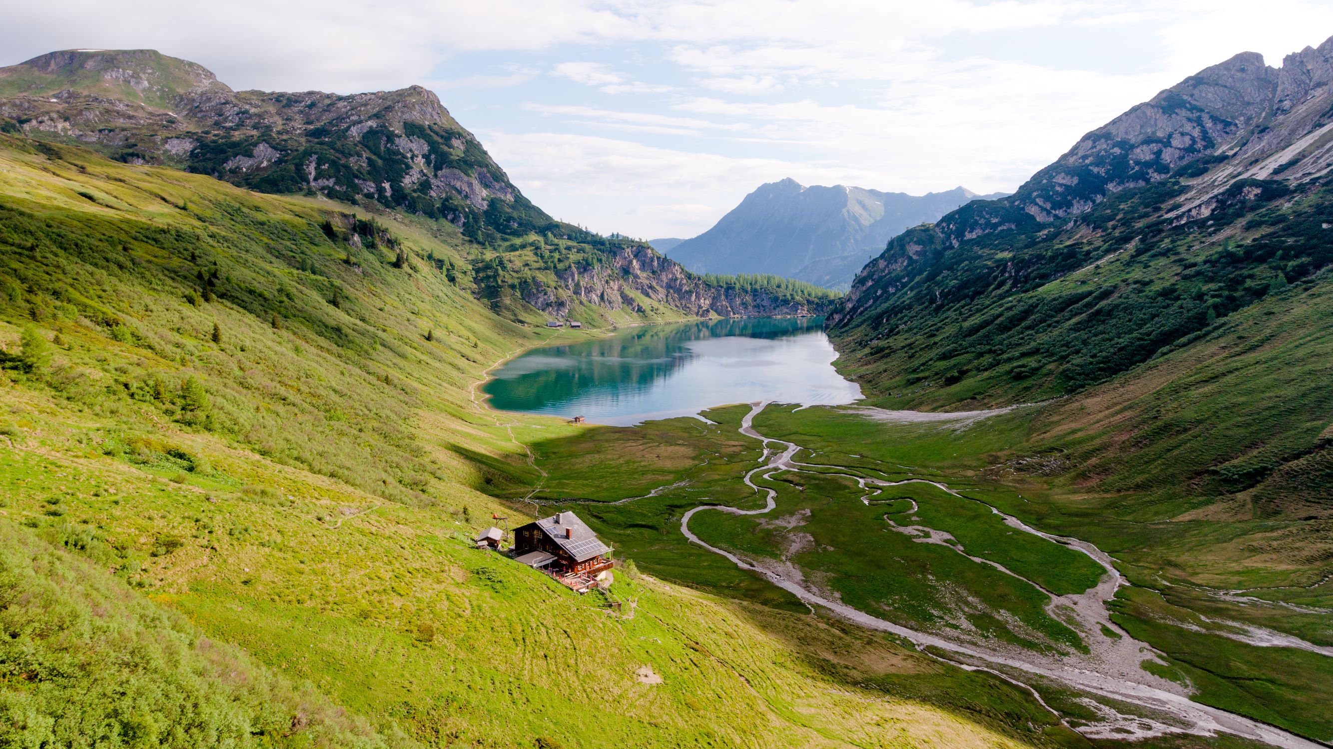 Bergpanorama im Sommer - saftige grüne Wiese - See im Hintergrund - kleine Hütte in den Bergen